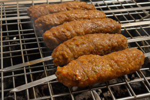 Grilled kofta kebabs being cooked over charcoal on a metal grill, showing raw seasoned meat skewers.